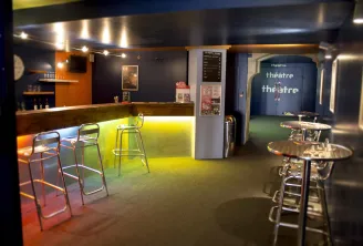 Interior of Théâtre Victoire in Bordeaux featuring a modern bar with yellow neon lighting, high stools, round tables, and the theatre entrance in the background