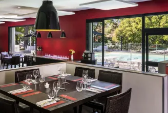 Modern restaurant featuring vibrant red walls, elegantly set black dining tables with glassware and place settings, dark pendant light fixture, and floor-to-ceiling windows overlooking a swimming pool and outdoor lounge area.