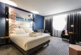 Modern hotel room at Mercure Bordeaux Centre featuring a white bed, headboard with Bordeaux cityscape illustration, dark blue walls, grey curtains, and a seating area with armchair.