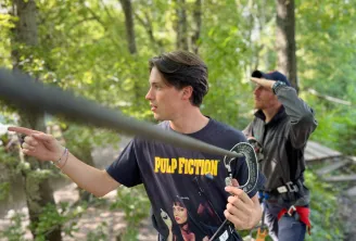 A young man wearing a Pulp Fiction t-shirt points ahead in a wooded area while participating in a treetop adventure park activity, with another participant visible in the background.