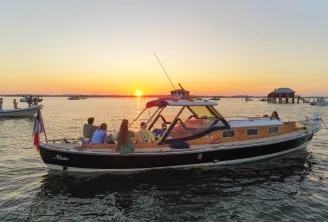 The Altaïr pinasse, a traditional orange and black Bordeaux boat, carries passengers during sunset on the Gironde estuary, with other vessels and oyster huts visible in the background.