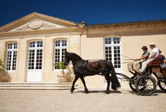 A black horse-drawn cart with three passengers in front of the classical facade of Château Desmirail in Bordeaux, under clear blue sky