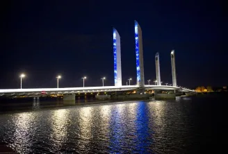 Bordeaux's Jacques Chaban Delmas Bridge lit up at night, featuring four blue illuminated vertical pylons reflected in the Garonne River