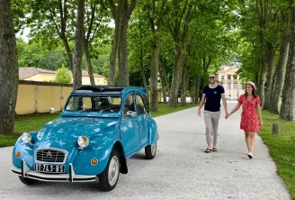 A turquoise Citroën 2CV with an open roof parked under a tree-lined avenue in Bordeaux, with a couple walking nearby and historic buildings in the background.