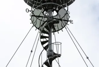 Bollée wind turbine at Beauval Estate in Bordeaux, low-angle view showing the metal structure with black helical blades, central mast and tension cables against a cloudy sky.
