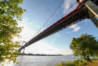 The Aquitaine Bridge, a large red and white suspension bridge spanning the Garonne River in Bordeaux, framed by green riverbank vegetation.