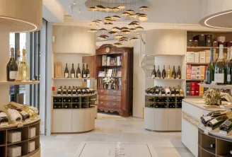 Elegant interior of a wine shop in Bordeaux featuring wine bottle displays on light wood and white shelving, a golden chandelier pendant light, and a vintage wooden cabinet in the center.