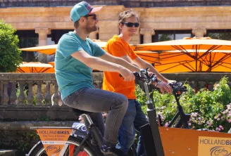 Two people riding an orange Holland Bikes rental bicycle in Bordeaux, with orange umbrellas and flowers in the background