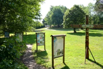Information boards and educational panels along a path in a lush park at Moulin Bidon site in Bordeaux, with trees and a wooden cross visible.