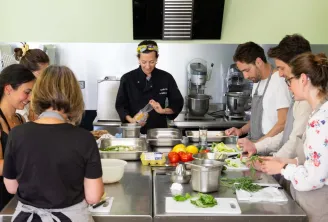 A cooking instructor in black demonstrates a culinary technique to a group of participants during a hands-on cooking class in Bordeaux. Students prepare fresh vegetables around a professional workstation equipped with electric mixers and kitchen tools.