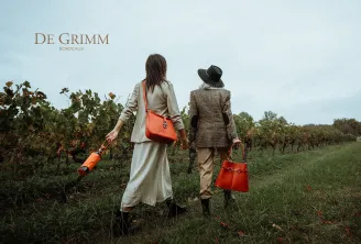 Two stylish women walk through Bordeaux vineyards, carrying red accessories and a wine bottle, embodying the luxury and wine heritage of the region.