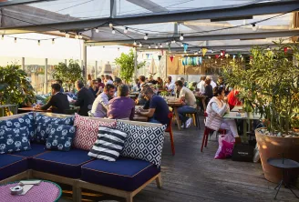 Covered rooftop terrace of Mama Shelter restaurant in Bordeaux with diners seated at tables, blue sofas with colorful cushions, green plants, string lights and decorative bunting.