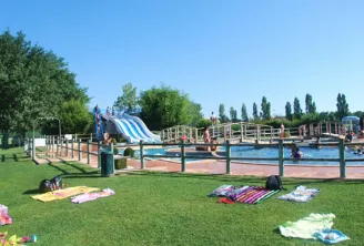 Pinsan swimming pool in Eysines featuring blue water slides, wooden bridges, swimming basins, green lawns, and visitors relaxing on towels.