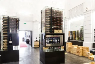 Contemporary interior of Max Bordeaux Wine Gallery & Cellar featuring illuminated wine displays with bottles in glass cases, stacked wooden wine crates, and tasting equipment in a bright, minimalist space.