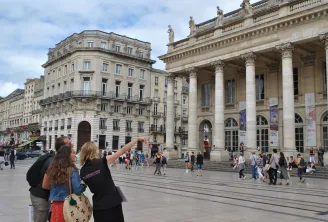 Two tourists admiring the Place de la Comédie in Bordeaux, with the majestic Grand Theatre and its impressive columns in the background, surrounded by visitors and Haussmann-style buildings.