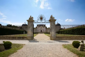 Overview of Château de Malle in Bordeaux, featuring ornate stone gateways with sculptures, wrought iron fencing, gravel drive, and a classical mansion with mansard roof.