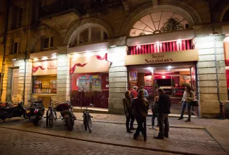 Night view of the Théâtre des Salinières in Bordeaux, featuring its distinctive red entrance, parked bicycles and scooters, and pedestrians on the cobblestone plaza.