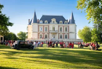 Château Lamothe Bergeron in Bordeaux, a grand neo-Renaissance castle with white stone and red brick turrets, hosting an outdoor reception with numerous guests dressed in red on the lawn under clear skies.