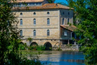 Porchères Mill in Bordeaux, an 18th-century stone building beside the Garonne River, featuring regular facades with arched windows and a stone arch spanning the water, surrounded by lush green vegetation.