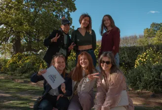 Six young people smiling and posing in a park in Bordeaux, with green trees and yellow flowers in the background.