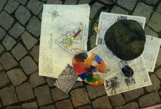 On a stone pavement, artist's documents and tools arranged: architectural sketch with compass, colorful paint palette, newspapers, black mirror, camera and creative supplies.