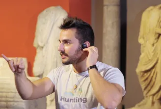 A man in a beige Hunting t-shirt talks on the phone in a museum exhibition, surrounded by ancient sculptures and orange walls.