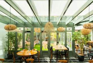 Restaurant dining room with large glass doors opening onto a terrace, featuring green exposed beams, natural rattan pendant lights, wooden tables and chairs, and indoor plants.