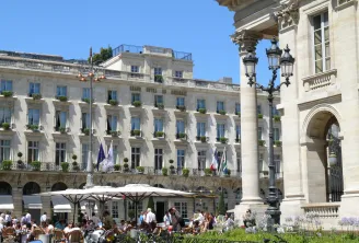 The Intercontinental Grand Hotel Bordeaux facade featuring its lively outdoor terrace, ornate balconies, Corinthian columns and flags under clear blue sky.