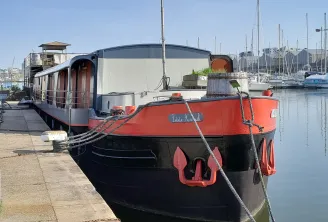 The Petite Annick houseboat moored at the quay in Bordeaux, featuring a red and black hull, surrounded by sailboats in the river port.