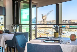 Restaurant terrace with set tables, blue chairs, and panoramic view of Bordeaux port with industrial cranes in the background