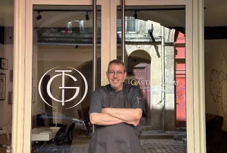 A chef smiles in front of The Gastronome Bordeaux storefront, a cooking school in Bordeaux, with arms crossed.