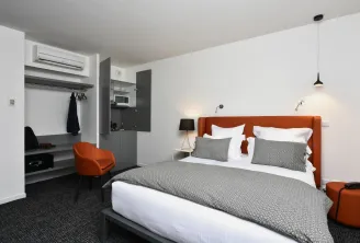 Modern hotel room at Ténéo Talence Espeleta in Bordeaux featuring a king-size bed with orange headboard, integrated work area, and gray and white furnishings.
