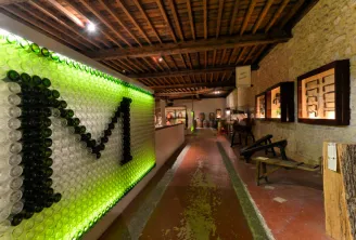 Interior of the Museum of Wine and Vine Crafts in Bordeaux, featuring an artistic installation of illuminated green and black bottles forming a graphic pattern on the left wall, with a terracotta exhibition corridor, display cases, and wooden ceiling beam
