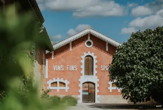Red brick façade of Maison Lillet in Bordeaux, featuring « VINS FINS » and « SPIR » inscriptions, white geometric ornaments, wooden doors, climbing ivy on the right side, cloudy blue sky.