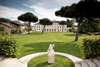 Malartic-Lagravière château in Bordeaux featuring its white neoclassical façade, expansive manicured lawn, a classical stone sculpture in the foreground, and umbrella pines framing the estate.