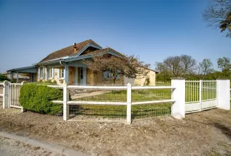 Colonial-style house with yellow and blue facade surrounded by white rail fencing, located in the Bastide de Siran district of Bordeaux, with trees and clear sky.