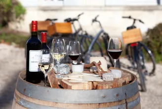 Wine and cheese tasting set up on a wooden barrel with red and white wine glasses, cheeses, and bicycles visible in the background on a paved courtyard.