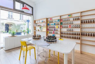 Bordeaux olfactory studio featuring a collection of wine bottles and luxury products displayed on wooden shelves, with a white table, yellow chairs, and large bay windows overlooking the city.