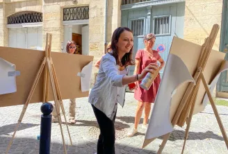 A woman paints on a cardboard easel in a paved courtyard in Bordeaux, surrounded by other participants in an outdoor art workshop.