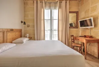 Elegant hotel room featuring a white bed, golden stone walls, light wood furniture, desk with chair, wall-mounted television, and tall windows with beige curtains in Bordeaux.