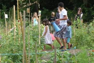A group of children in a vegetable garden at Parc Rivière in Bordeaux, where a young adult demonstrates watering plants with a blue watering can.