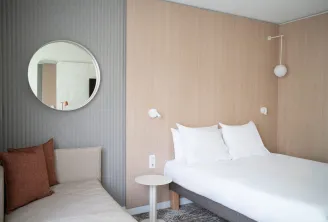 Modern hotel room at Novotel Bordeaux Mérignac featuring a double bed with white linens, minimalist furnishings, round mirror, and beige and grey tones.