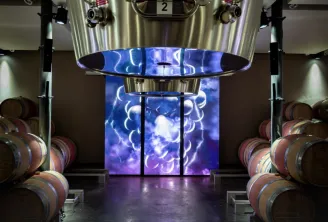 Modern winery facility at Château La Fleur de Boüard in Bordeaux, featuring oak barrels on either side and a large digital screen displaying an illuminated grape cluster pattern in blue and purple.