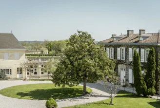 Aerial view of Château Prieuré-Lichine in Bordeaux featuring white stone buildings with tiled roofs, a large tree in the courtyard, gravel pathways, and manicured gardens with vineyards in the background.