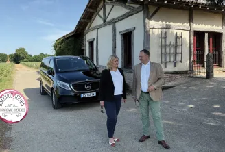 Two people standing in front of a traditional Bordeaux house with a black Mercedes vehicle, vineyards in the background, Bacchus Tours logo visible