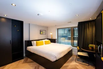 Modern aparthotel bedroom featuring a queen-size bed, black furniture, yellow accents, large windows overlooking Bordeaux, and contemporary amenities.