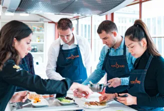 Four people wearing blue Côté Cours aprons participate in a cooking class in a modern kitchen with large windows overlooking Bordeaux. They are preparing dishes together at a work counter.