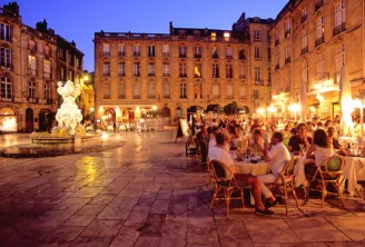 Evening view of Place de la Bourse in Bordeaux with outdoor restaurant seating, an illuminated fountain in the center, and historic golden stone buildings