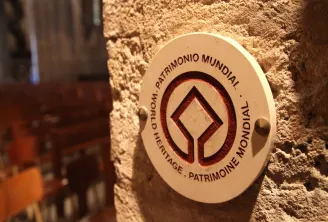 Circular UNESCO World Heritage plaque mounted on a stone wall, featuring a stylized diamond symbol and multilingual text indicating world heritage status.