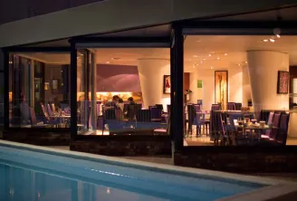 Night view of GourmetBar by Novotel Bordeaux Lac featuring an illuminated glass façade overlooking a swimming pool, with elegantly set dining tables visible inside